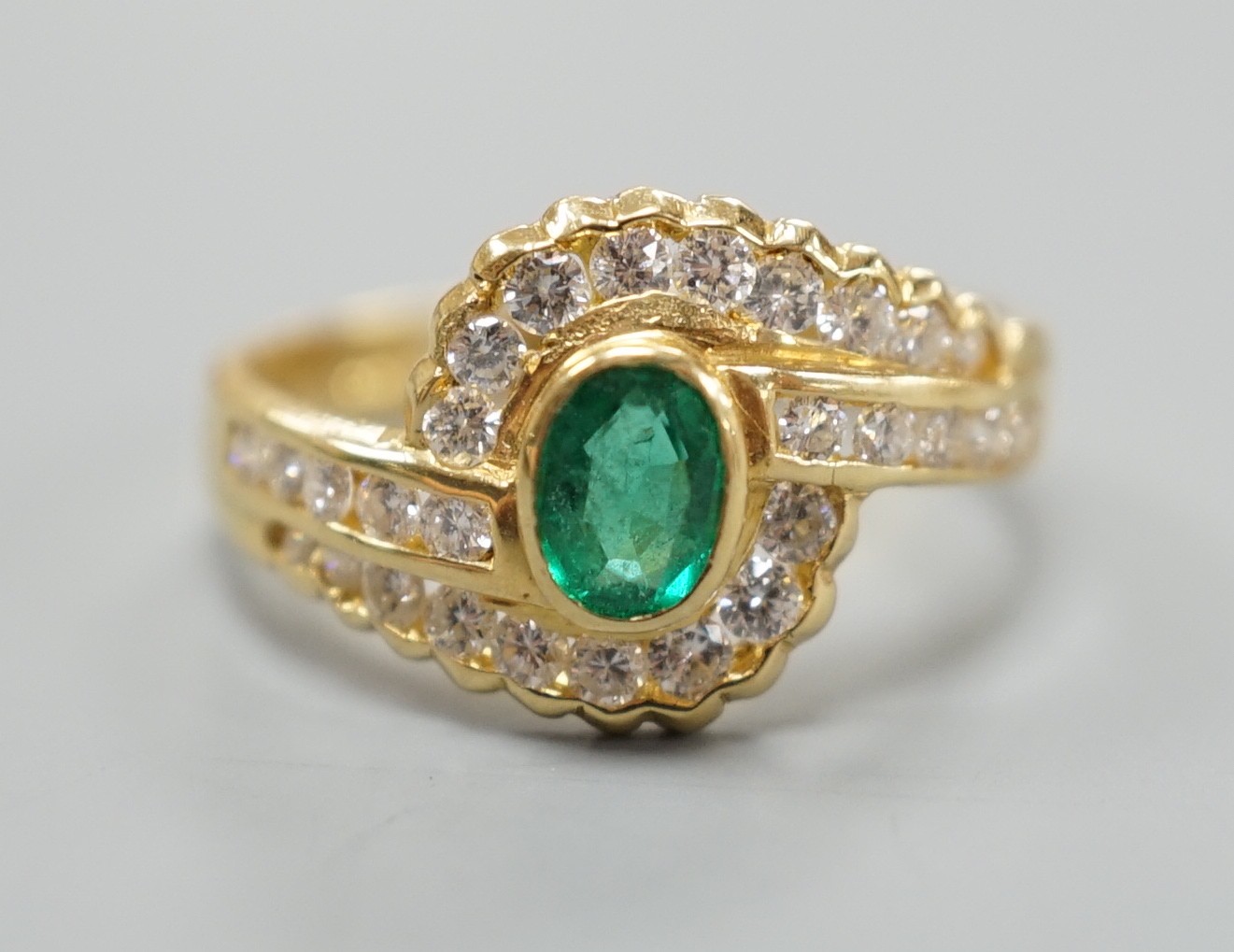 A modern 18ct gold, collet set oval cut emerald and diamond cluster set crossover ring, size N, gross weight 3.5 grams.